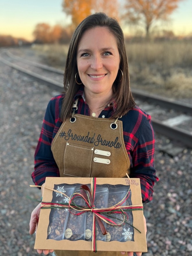 A woman smiling at the camera while holding a holiday box of assorted granola wrapped with red and green twine.
